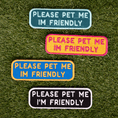 Load image into Gallery viewer, Four colorful "Please Pet Me I'm Friendly" patches on grass, featuring blue, pink, yellow, and black backgrounds.