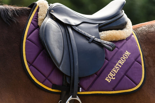 Dark Orchid Saddle Pad by Southlake Paws featuring a quilted design, gold trim, and a saddle placed on a horse.