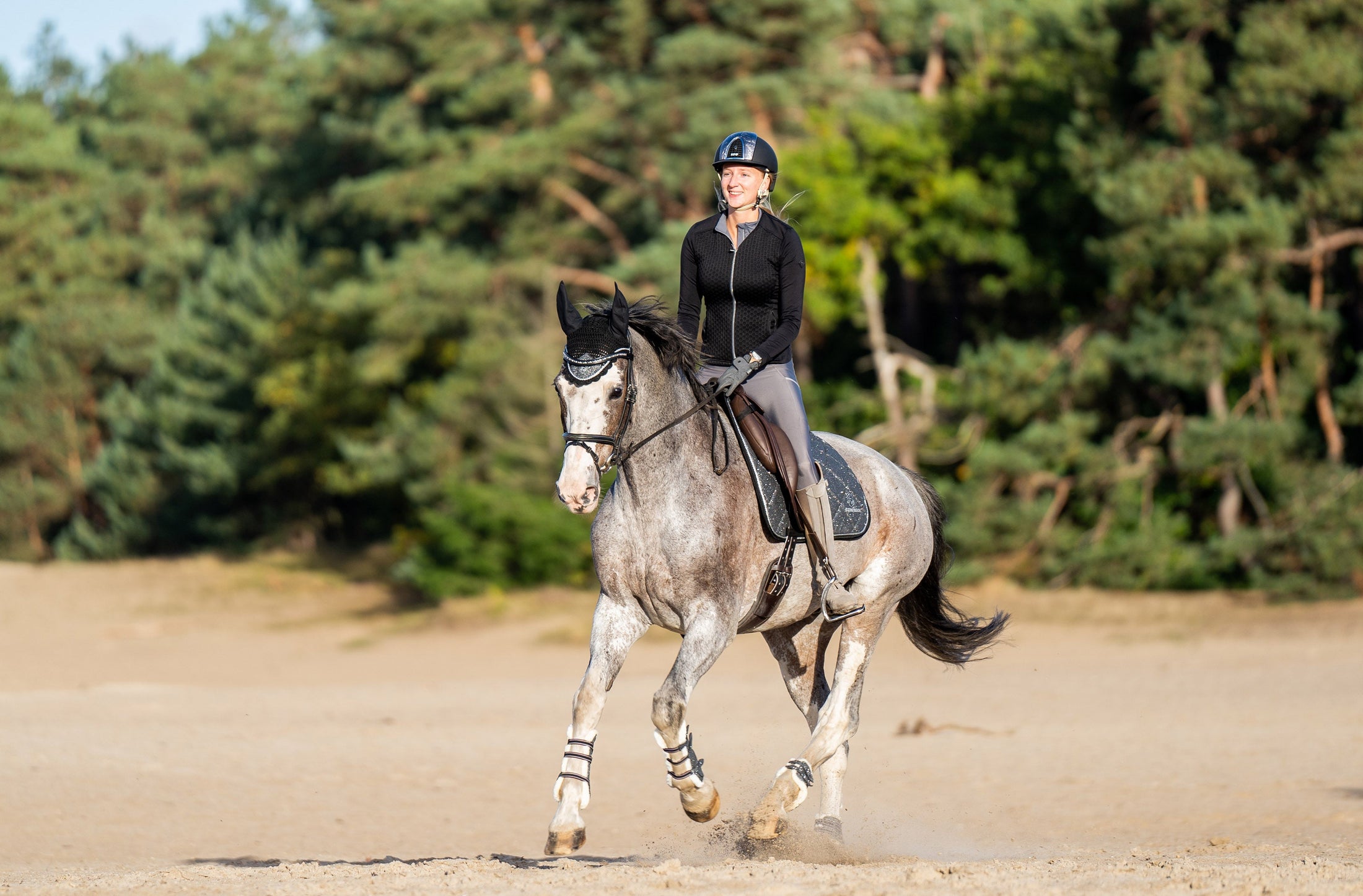 A rider in black attire on a grey horse, showcasing a Stardust Grey Saddle Pad in a sandy outdoor setting.
