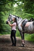 Load image into Gallery viewer, A rider in a pink shirt stands beside a gray horse wearing a powder pink suede saddle pad set in a wooded area.
