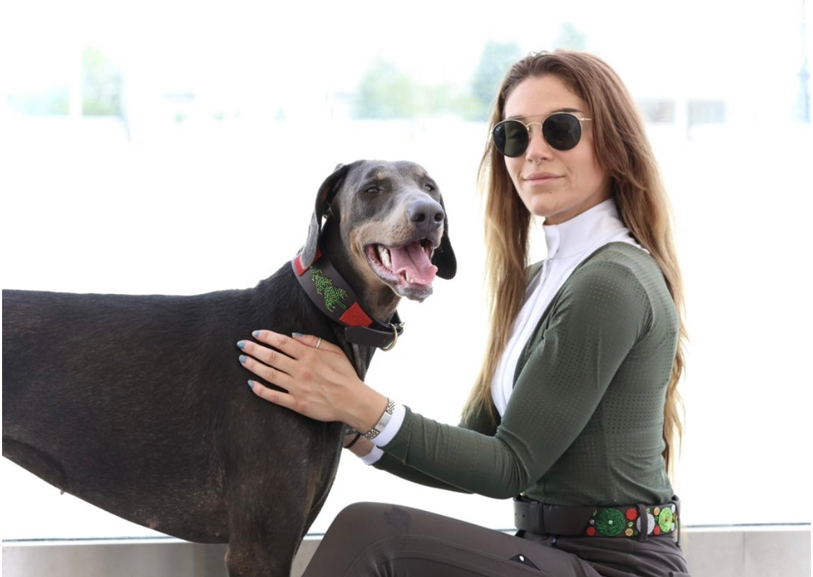 A woman wearing sunglasses sits beside a happy dog, showcasing a festive beaded belt from Southlake Paws.