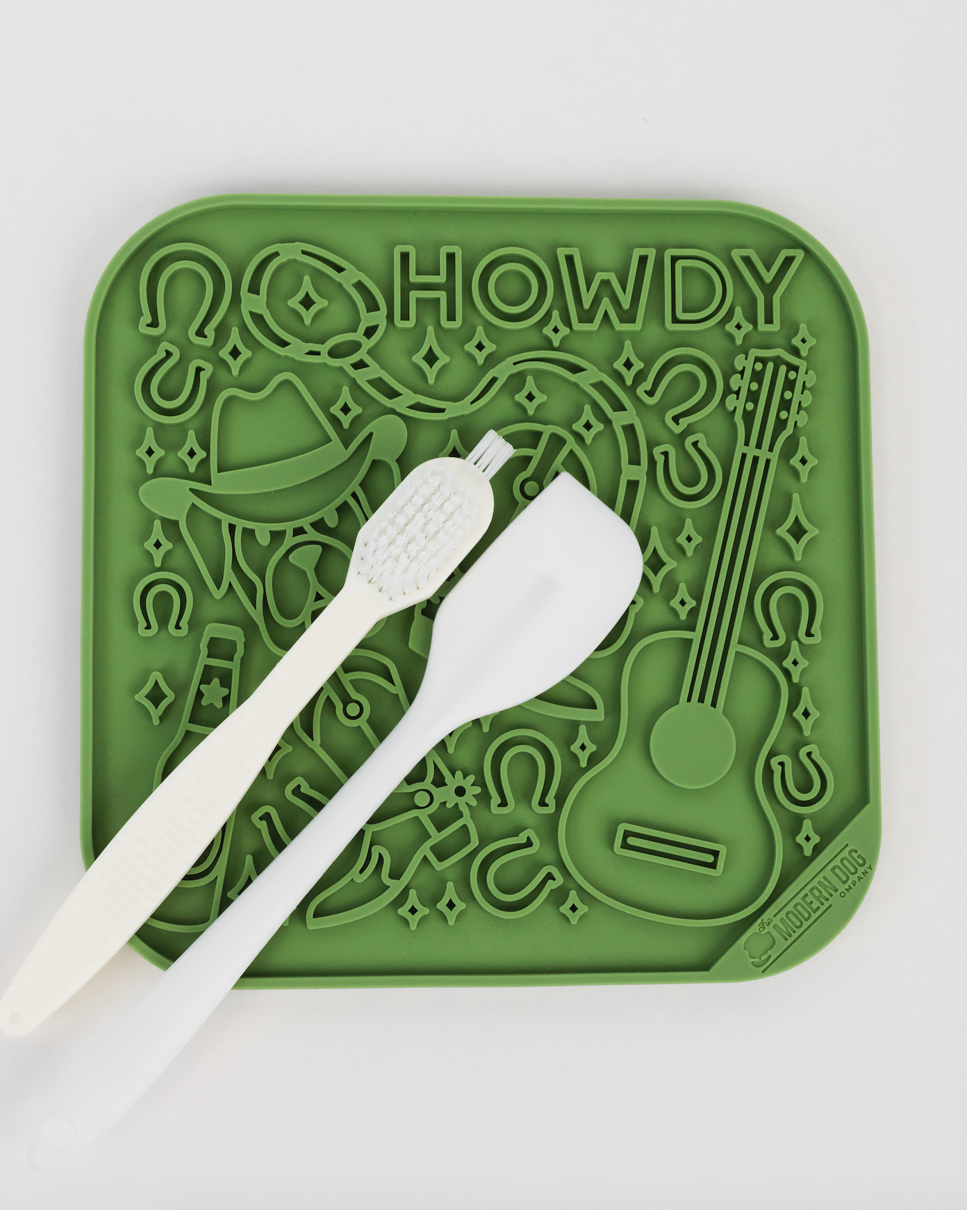 Green silicone lick mat featuring "Howdy" design with a guitar and cowboy elements, accompanied by a toothbrush and spatula.