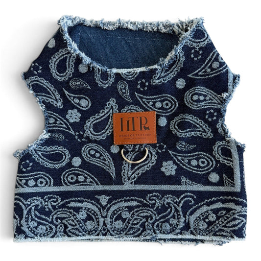 A small dog harness made from dark blue denim with a light blue paisley pattern, featuring a brown leather brand patch and a metal ring attachment.