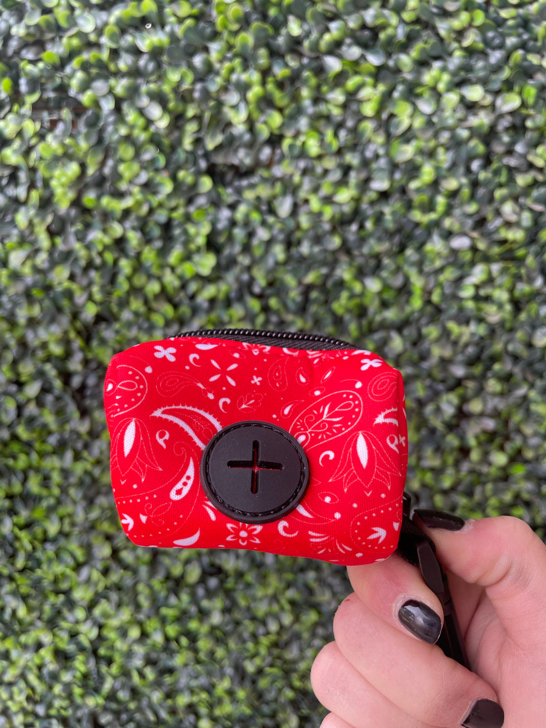 Red Paisley Poop Bag Holder by The Modern Dog Company, featuring a zippered design and a button closure, held against a green