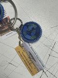 Load image into Gallery viewer, Beaded keychain from Southlake Paws featuring a circular blue design, attached to a keyring with a tag about its Kenyan origi
