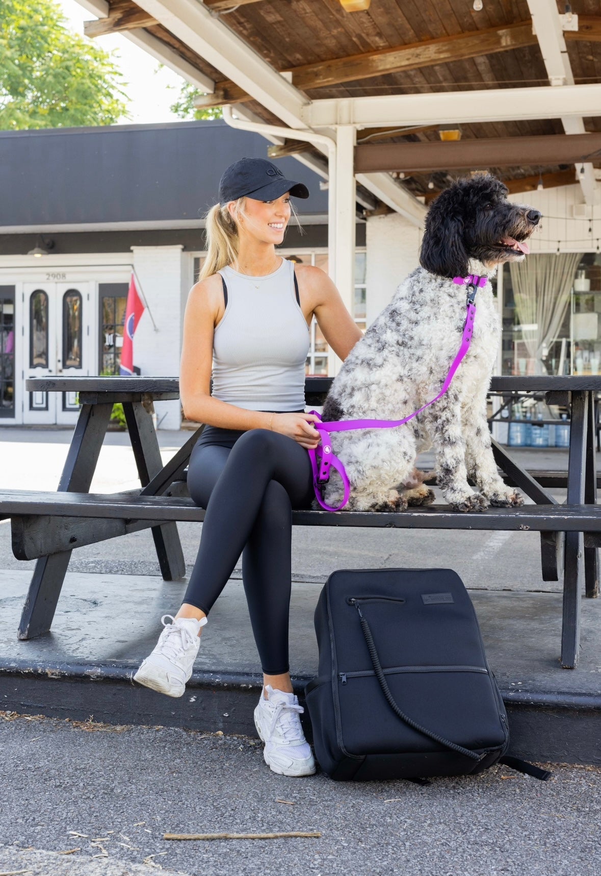 Woman in athletic wear sits on a bench with a dog wearing an electric fuchsia collar, next to a black backpack.