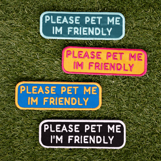 Four colorful "Please Pet Me I'm Friendly" patches on grass, featuring blue, pink, yellow, and black backgrounds.